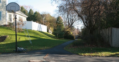 This figure is an image showing a pedestrian path used by students to reach their school bus pickup point. There is a basketball hoop at the end of the nearby street.